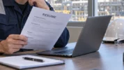 Inspection technician reviewing a resume at a desk with safety gear and refinery piping visible through the window.